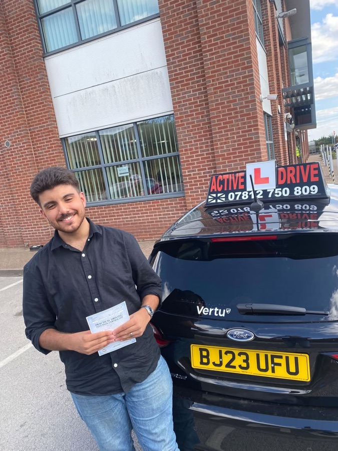 A real Active Drive student smiling broadly and holding his pass certificate in front of the Active Drive car, outside a brick office building