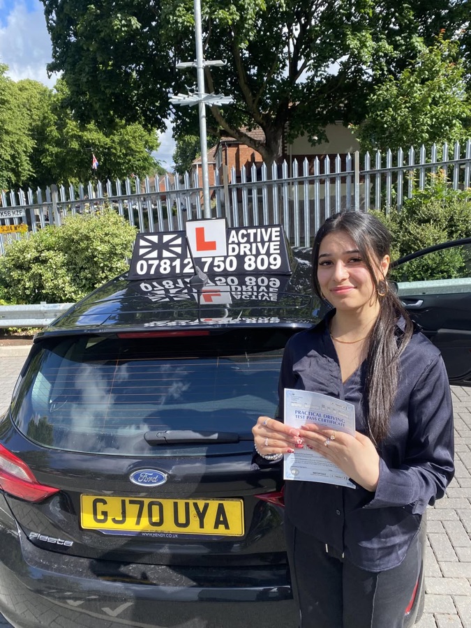 A real Active Drive student holding her pass certificate beside the Active Drive estate car, with metal railings and a tree behind her