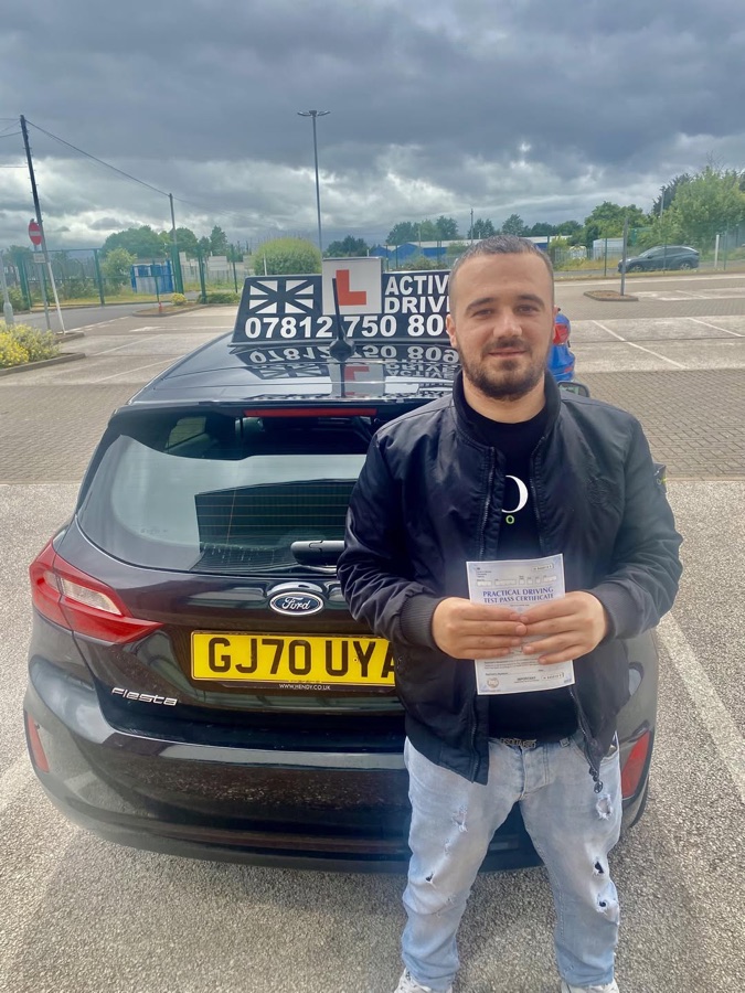 A real Active Drive student in a dark bomber jacket holding his pass certificate beside the Active Drive car under a cloudy sky