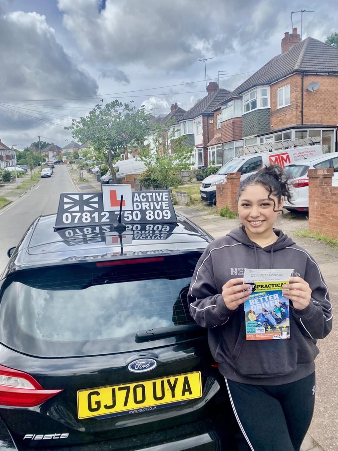 A real Active Drive student holding her pass certificate and a new-driver magazine on a residential street, beside the Active Drive car