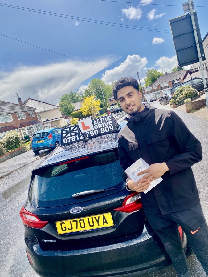 A real Active Drive student in a dark jacket holding his pass certificate beside the Active Drive car on a rainy residential street
