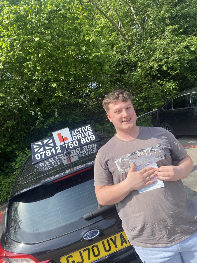 A real Active Drive student holding his pass certificate beside the Active Drive car, with trees and another car behind him